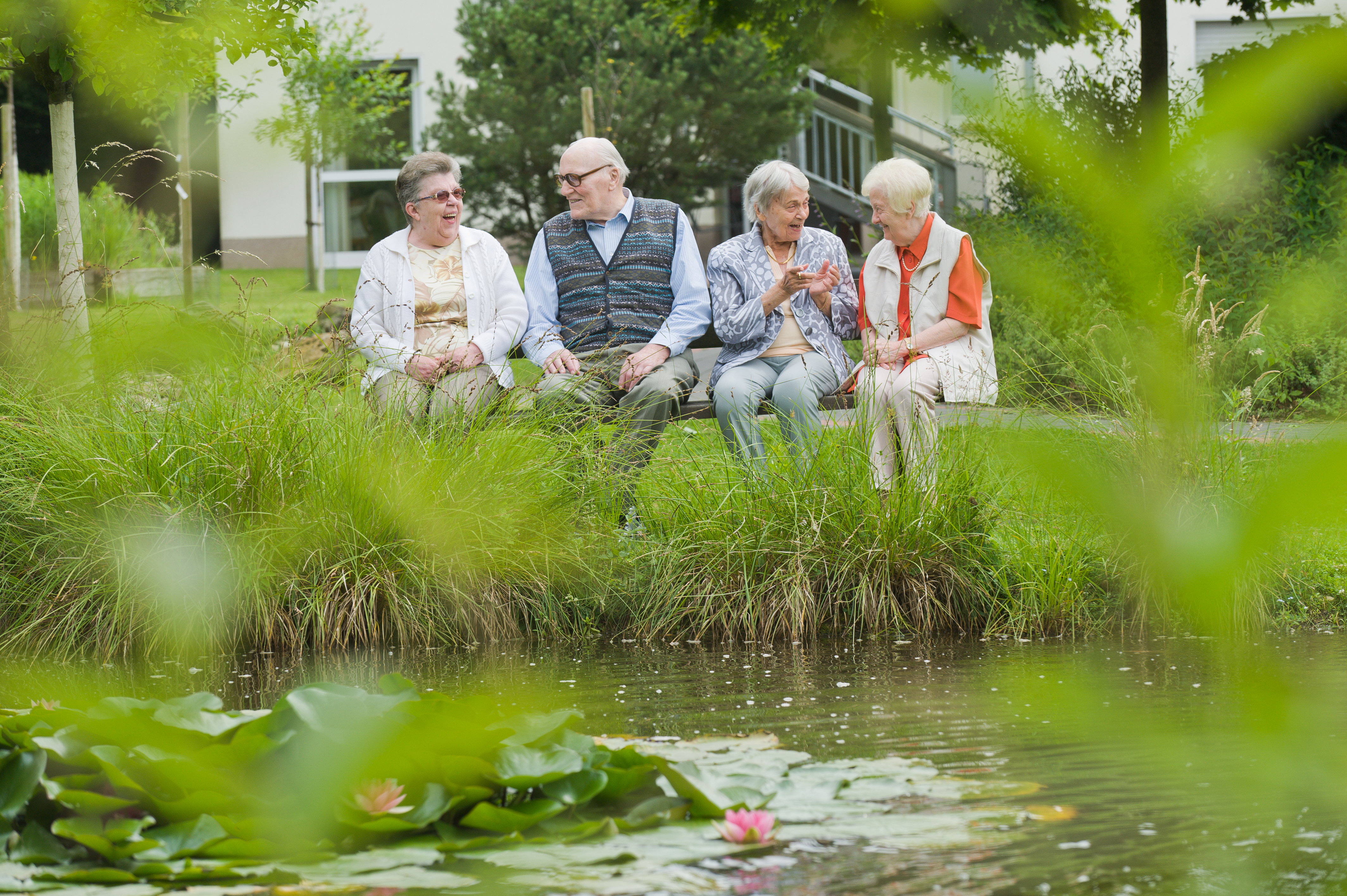 Vier Bewohner eines Altenheims sitzen an einem See auf einer Bank und unterhalten sich.