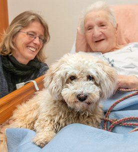 Eine Bewohnerin aus einem Seniorenzentrum kuschelt mit einem Hund in Ihrem Pflegebett. Eine Pflegefachfrau ist bei ihr und lächelt.