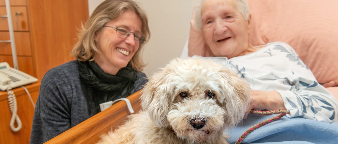 Eine Bewohnerin aus einem Seniorenzentrum kuschelt mit einem Hund in Ihrem Pflegebett. Eine Pflegefachfrau ist bei ihr und lächelt. 190404_DW_066.JPG
