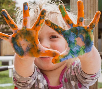 Ein lächelndes kleines Mädchen zeigt stolz seine bunten Hände, die mit Fingerfarbe bepinselt wurden.
