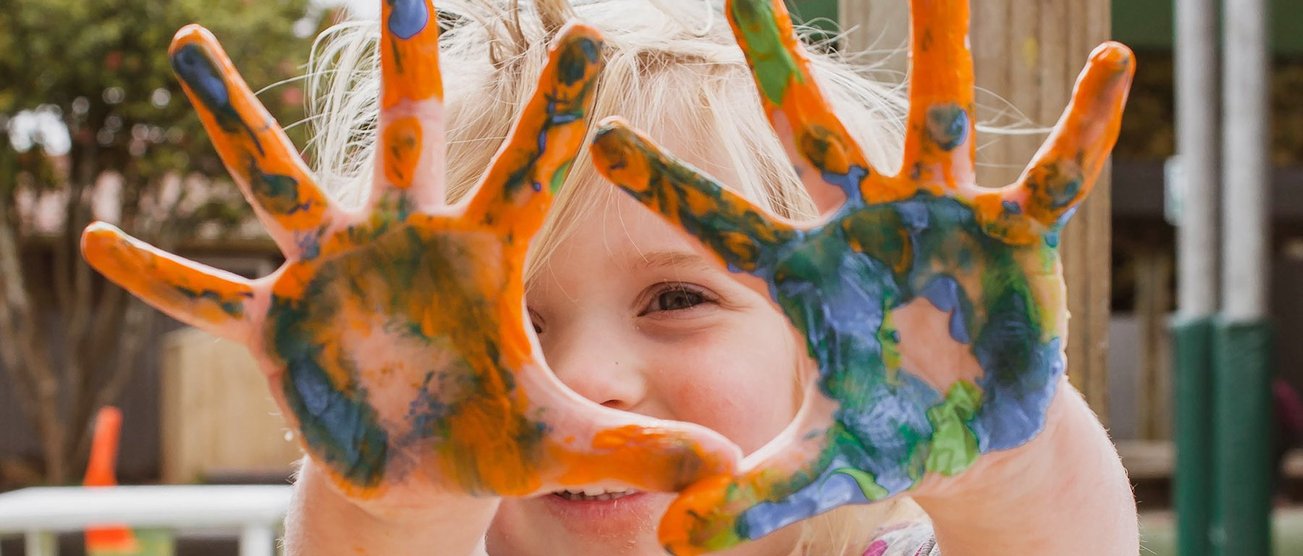 Ein lächelndes kleines Mädchen zeigt stolz seine bunten Hände, die mit Fingerfarbe bepinselt wurden. EDE.jpg