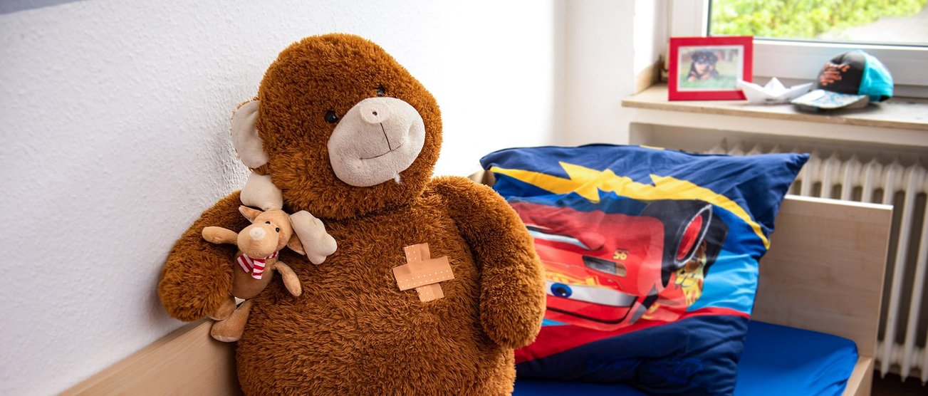 Ein gemütliches Kinderzimmer mit einem Bett. In dem Bett sitzt ein großer lächelnder Teddybär. Inobhutnahme_Kinder.JPG