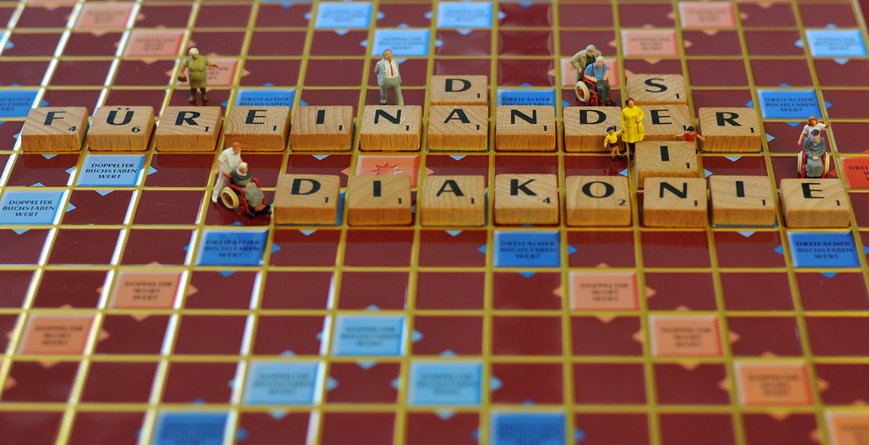 Ein Scrabble-Brett mit den gelegten Wörtern "Füreinander das sein Diakonie". Um die Holzplättchen sind kleine Figuren gestellt, die Pflege symbolisieren.