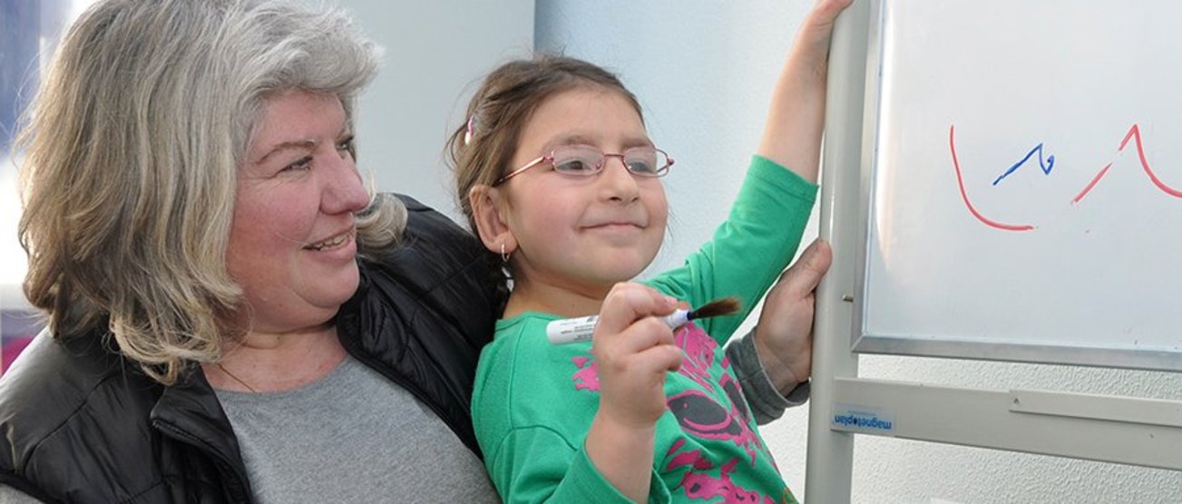 Eine Pädagogin hält ein aufgewecktes Kind in die Höhe so das, dass junge Mädchen mit einem Stift an eine Tafel schreiben kann. _Schulbegleitung_hp.jpg