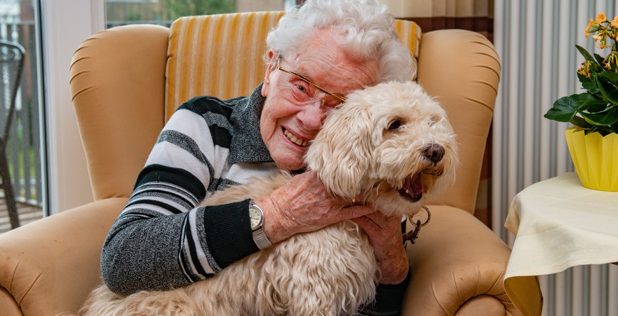 Eine Bewohnerin aus einem Seniorenzentrum kuschelt mit einem Hund auf Ihrem Sessel.