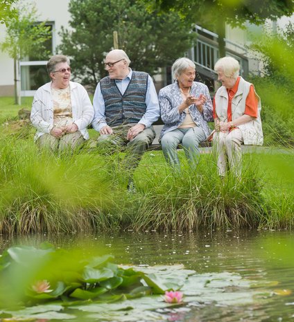 Vier Bewohner eines Altenheims sitzen an einem See auf einer Bank und unterhalten sich.