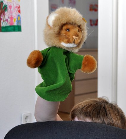 Ein Kind versteckt sich hinter einem Stuhl und hält eine Löwen-Handpuppe hoch.