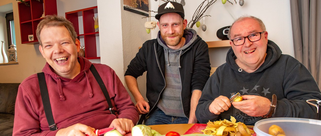 Drei Männer sitzen fröhlich an einem Küchentisch und schälen Gemüse für das Mittagessen. 190404_DW_072.JPG