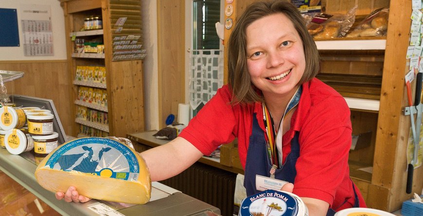 Eine lächelnde Verkäuferin eines Hofladens zeigt unterschiedliche Käsesorten.
