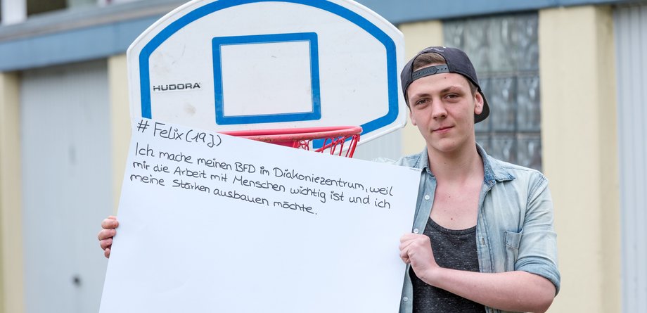 Ein junger Mitarbeiter des Diakonischen Werks steht draußen vor einem Basketballkorb und hält ein Schild hoch.
