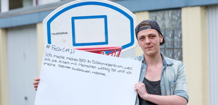 Ein junger Mitarbeiter des Diakonischen Werks steht draußen vor einem Basketballkorb und hält ein Schild hoch.
